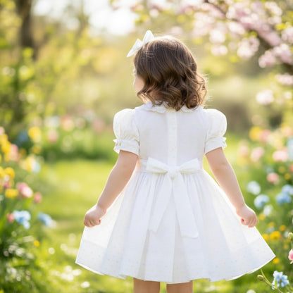 Back view of a toddler girl in a white hand-smocked dress with pink flower basket embroidery and a bow in a garden.