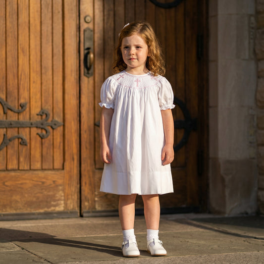 Young girl wearing a white smocked christening dress with pink crosses, standing by wooden church doors.